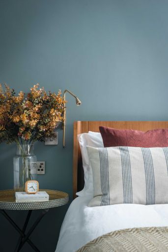 Kohtalo. Close-up of bed. Striped pillow, blue painted wall. Vase of flowers on bedside table